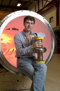 Alex Romanenko, sitting on the edge of a large cryogenic vessel, holds one of the superconducting RF cavities made of niobium. Photo: Fermilab (Click on image for larger version)