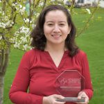 Jeny Teheran holds her SHPE award, which is made of transparent glass with words etched on it. She wears a red shirt and the background is a bright green field and blossoming tree or shrub.