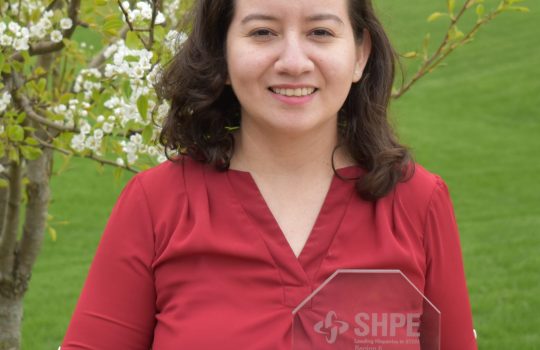 Jeny Teheran holds her SHPE award, which is made of transparent glass with words etched on it. She wears a red shirt and the background is a bright green field and blossoming tree or shrub.