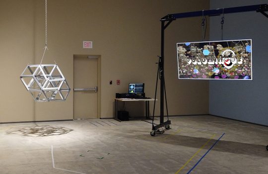 A large silver hive-like object that looks like it's made from an erector set hangs from the ceiling. Its shadow on the floor below. A door with an exit sign is in the center of the photo. A projection screen with an illustration that reads "voluminous" is on the right side of the photo.