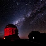 A starry night sky with purple diagonal stripe from lower left to upper right corner above an observatory lit up in bright red. A shadow of a building or facility is in the lower right corner.