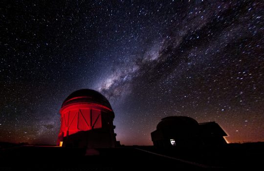 A starry night sky with purple diagonal stripe from lower left to upper right corner above an observatory lit up in bright red. A shadow of a building or facility is in the lower right corner.