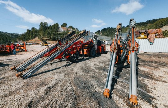 An orange and silver drill rig (a tractor-like apparatus with two parallel arms that reach above the cab and then make a steep diagonal to the ground) and several red and silver drill rigs sit in the foreground of a silty construction site. Other equipment is in the midground and hills filled with evergreens and blue sky above in the background.