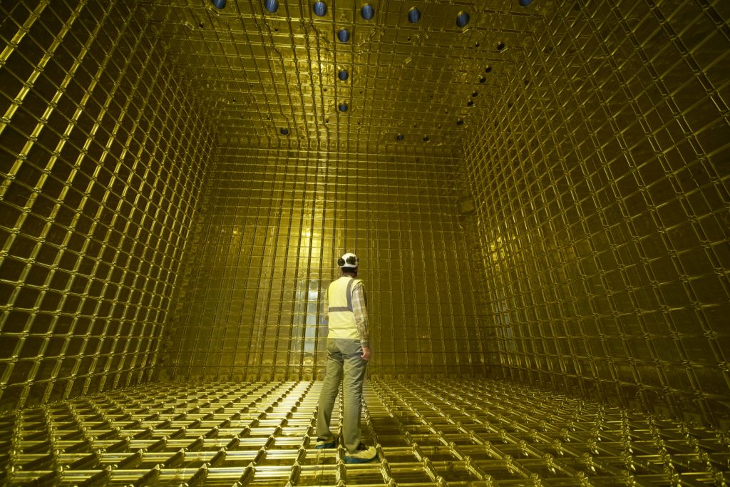 A person wearing a hardhat stands inside a bright yellow honeycomb-like box that appears to be two or three times their height.