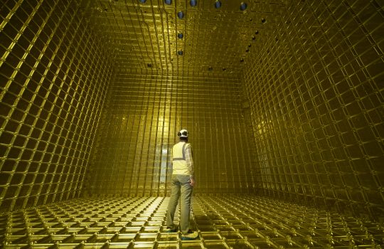 A person wearing a hardhat stands inside a bright yellow honeycomb-like box that appears to be two or three times their height.