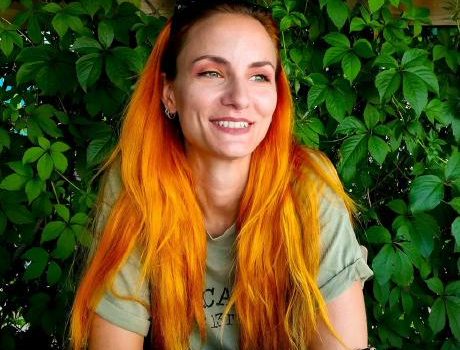 A photo of a woman with long, bright-orange hair, wearing sunglasses on top of her head and a light green T-shirt, smiling. Behind her, greenery.