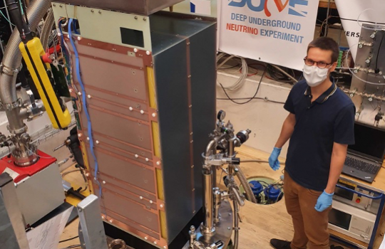 A large box-like structure sits on a round platform. Behind it, a banner that says "DUNE." To the right, a man with short hair wears glasses, a face mask, blue surgical gloves, a blue shirt and brown pants.