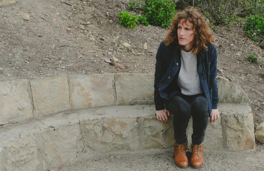 A woman with shoulder length brown hair sits on a stone wall.