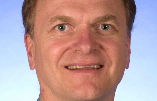 Headshot of a man with short auburn hair wearing a blue button-up smiles against in front of a gradient blue background.