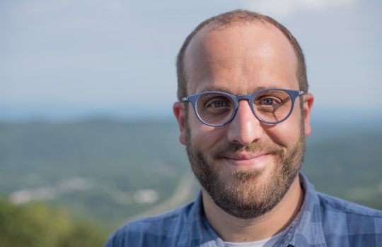 A man with close-cut brown hair and beard wears blue glasses and smiles at camera. In the background, blurred mountains, valley and sky.