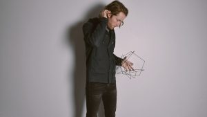 A man with brown chin-length hair in profile. He tucks his hair behind an ear with one hand and holds a small wire geometric sculpture in the other. He wears a dark outfit and is against a light gray wall.