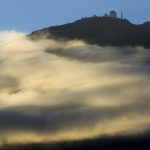 Photo of a dome-shaped building, likely an observatory, atop a mountain, which gold mist surrounds. Blue sky and silhouette of birds above.