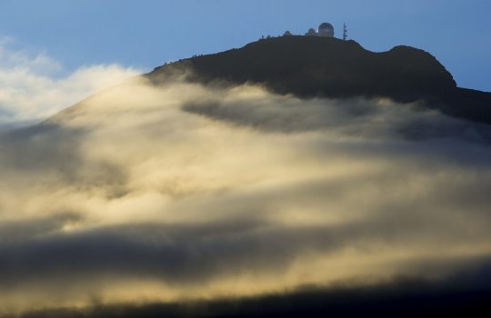 Photo of a dome-shaped building, likely an observatory, atop a mountain, which gold mist surrounds. Blue sky and silhouette of birds above.