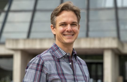 A photo of a man in a buttonup with short hair smiling in front of a concrete and glass building.