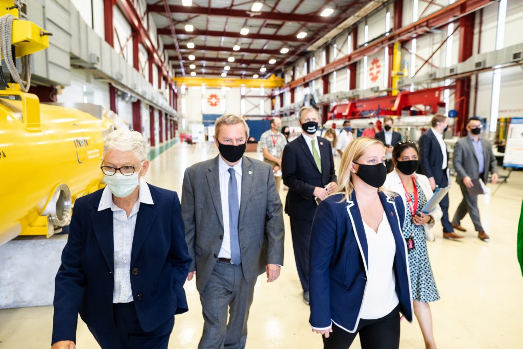Group of masked people in business attire walk through a high-ceiling building.