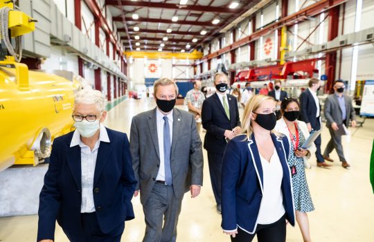 Group of masked people in business attire walk through a high-ceiling building.