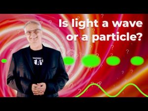 A man with gray hair, glasses and a black blazer smiles. To the right of him, the question "Is light a wave or a particle?"