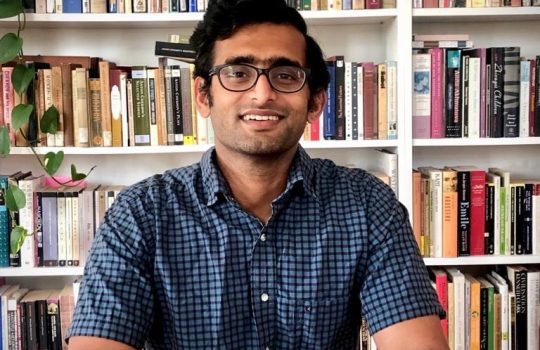 A photo portrait of a man with dark hair, glasses and a short-sleeve button-up shirt smiling, hands resting on a table in front of him, in front of a bookshelf.