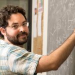 Scientist and 2021 URA Early Career Award winner stands at a chalkboard working on equations.