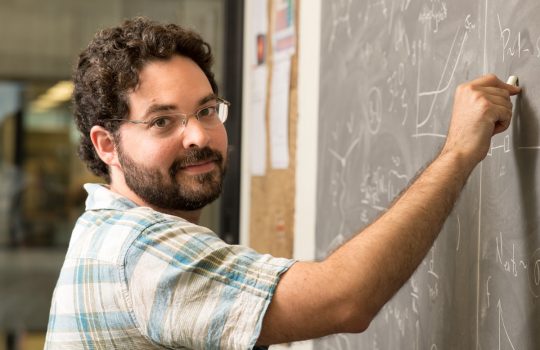 Scientist and 2021 URA Early Career Award winner stands at a chalkboard working on equations.