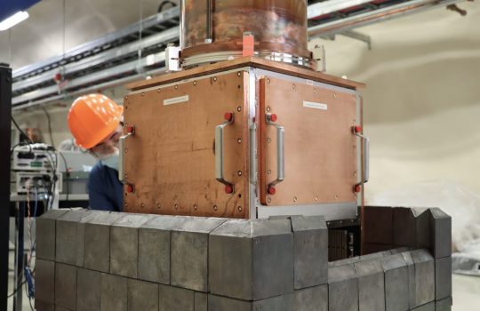 A person in PPE peers out from behind a large copper cube with doors with slate-colored blocks around its base.