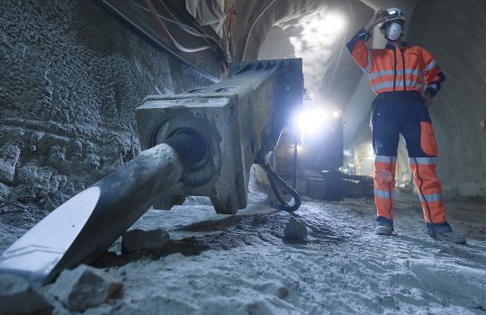 A person in a face mask and construction clothing adjusts the light on their hardhat stands in a gray tunnel near equipment. Behind, a large light shines.