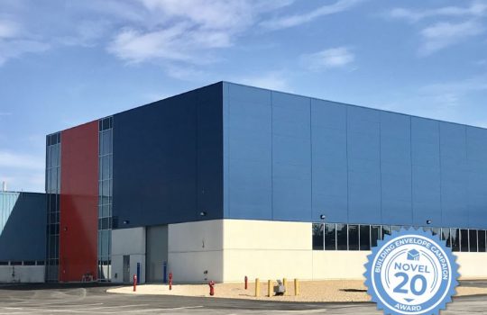 Photo of one end of a large rectangular building that's concrete, blue, gray and red with blue sky above. An award logo is superimposed in the lower right corner. It reads: "Building Envelope Campaign Novel 20 Award."