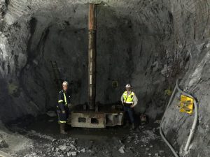 Two men stand underground next to a very large reaming drill bit