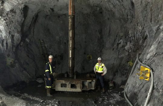 Two men stand underground next to a very large reaming drill bit