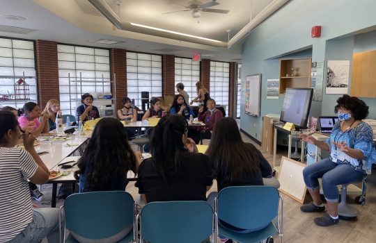 Girls around a U-shaped table listen intently as a speaker presents a talk.