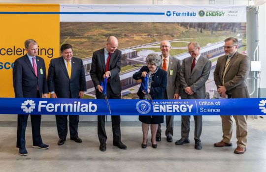 Deputy Secretary of Energy David Turk and Fermilab Director Lia Merminga cut a large ribbon with oversized ceremonial scissors while other people look on