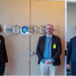 Three man stand in front of a door with a large metal tag that reads 'SQMS'