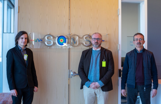 Three man stand in front of a door with a large metal tag that reads 'SQMS'