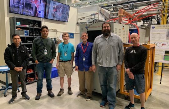 Six men stand in front of a prototype 40-kilowatt amplifier for PIP-II