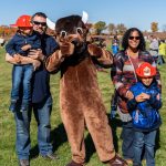 A bison mascot gives thumbs up to the camera while standing in between a family of four; the two boy children wear a red plastic firefighter helmet.
