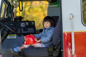 A toddler girl sits inside a fire engine holding a red firefighter plastic hardhat.