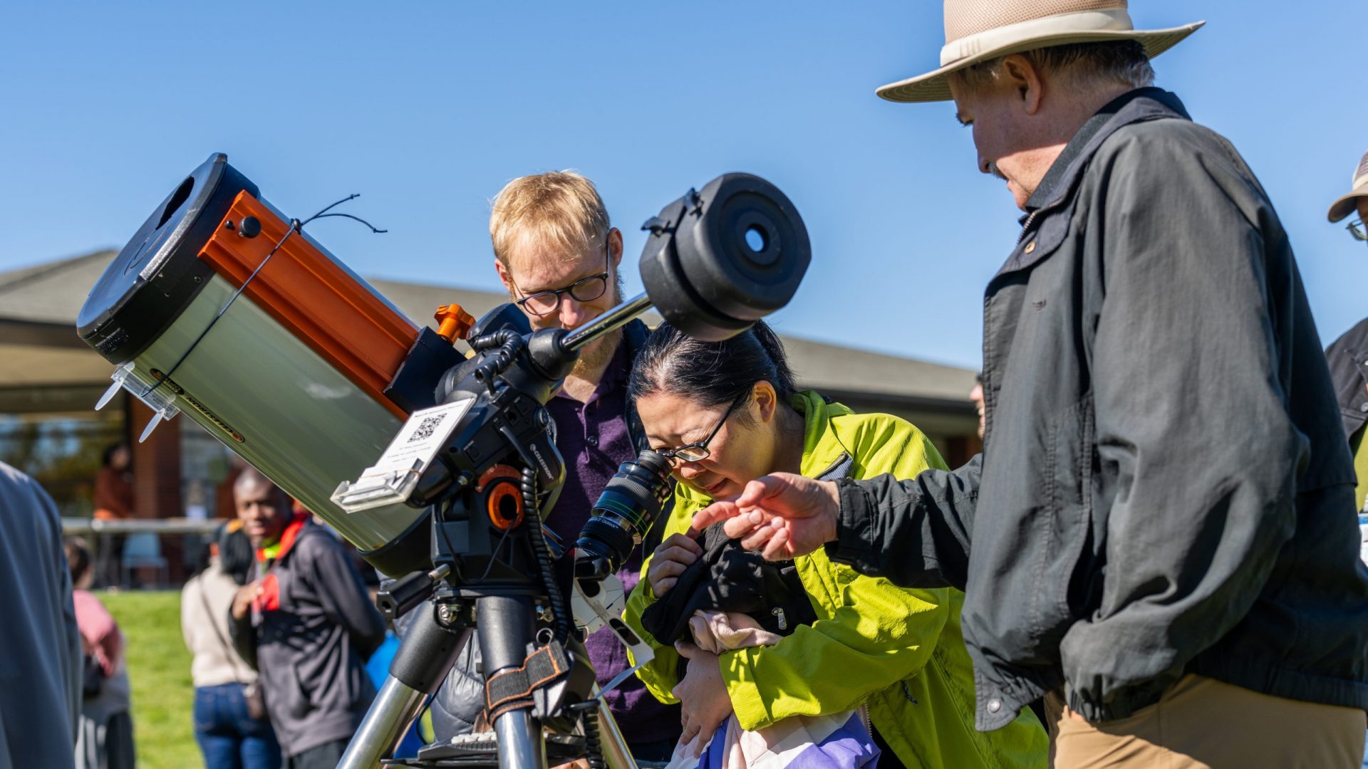 A woman with eyeglasses uses a telescope with sun filter, while a man with a hat gives her instructions on how to identify sunspots.