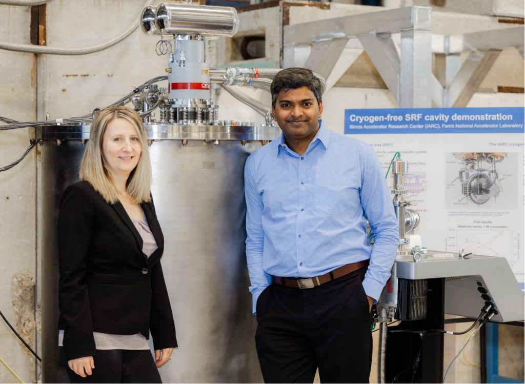 Two people stand next to the conduction-cooled cryostat that houses superconducting accelerator technology for industrial electron beam applications.