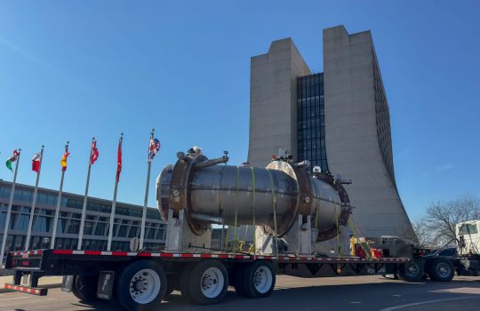 Truck with a long platform transporting a large curved apparatus drives in front of a tall building; international flags are visible on the left