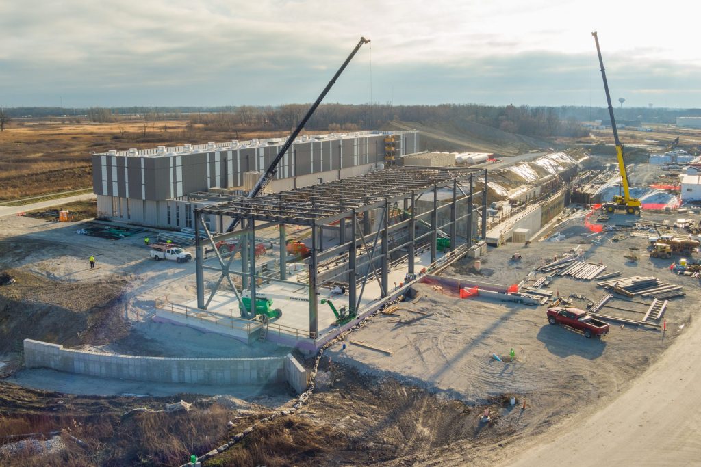 Construction site with two cranes lifting materials, a building under construction in the foreground, and a completed building in the background.