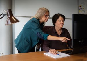 Two women collaborating on a project: one standing and pointing to a computer monitor while the other sits and listens attentively.