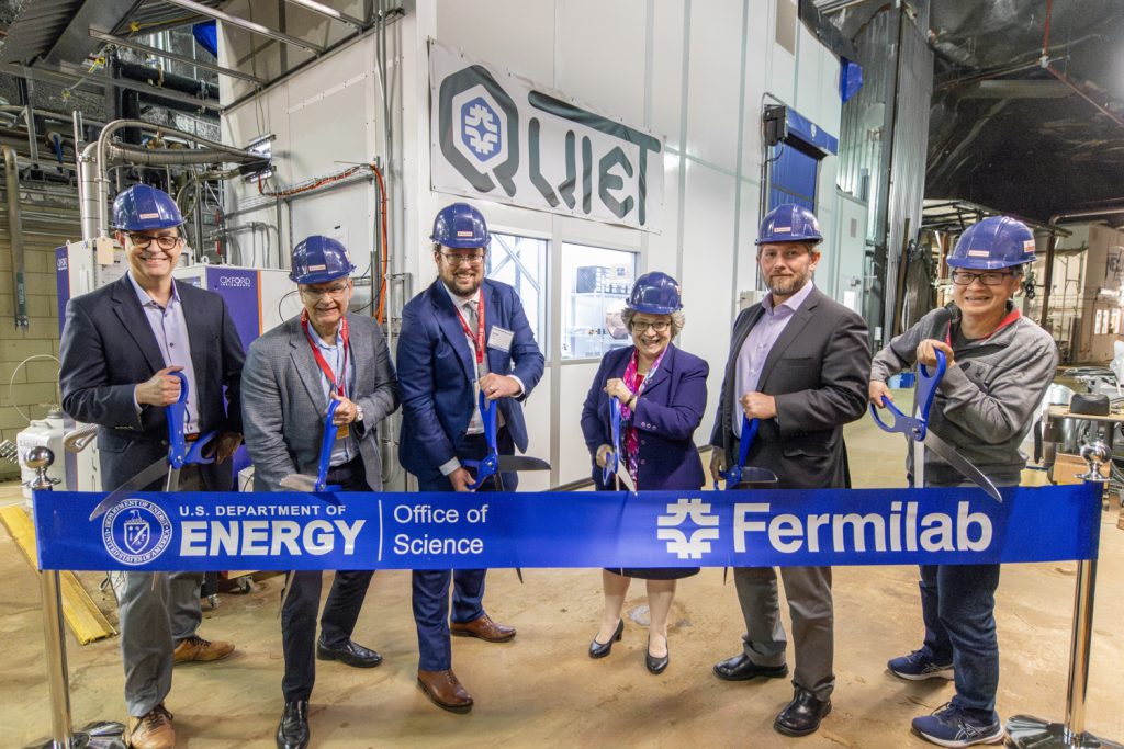 Six people wearing hard hats and holding scissors participate in a ribbon-cutting ceremony.