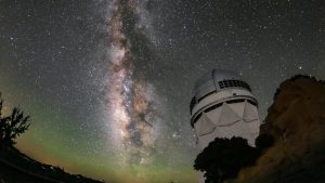 A white structure with a domed top on a rocky outcrop below a star-filled night sky. The Milky Way stretches vertically in a luminous band. Faint green glow tints the curved horizon.