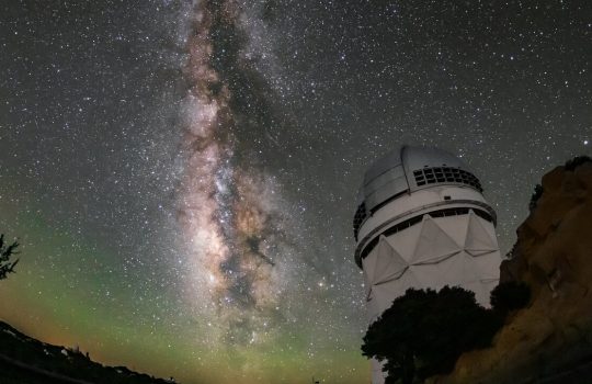 A white structure with a domed top on a rocky outcrop below a star-filled night sky. The Milky Way stretches vertically in a luminous band. Faint green glow tints the curved horizon.