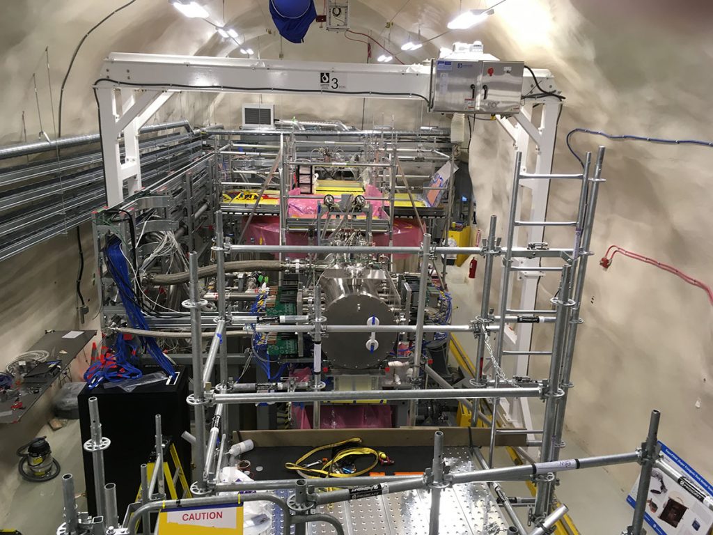 SuperCDMS is installed underground at SNOLAB. In the foreground is the end of a cylindrical vessel, the E-tank, which is the interface between the detector readout cables and the room-temperature electronics that digitize the detector signals. The yellow platform surrounds the SNOBOX cryostat that houses the detectors, and the pink plastic is covering the cylindrical shielding around the SNOBOX. The white gantry crane overhead is used for installation of heavy items. Credit: Lauren Hsu
