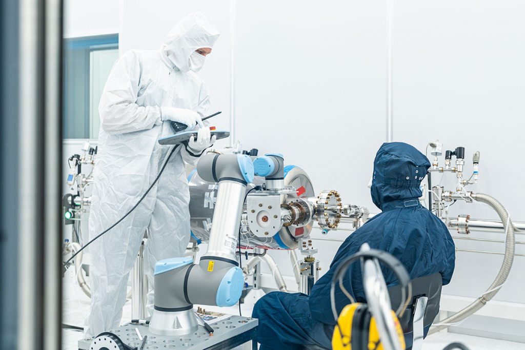 SRF engineer Tommaso Aiazzi and cleanroom technician Marco Battistoni work alongside a collaborative robot, or “cobot,” inside a clean room to install a high-power coupler on a cavity for Fermilab’s PIP-II accelerator. Credit: Ryan Postel and JJ Starr